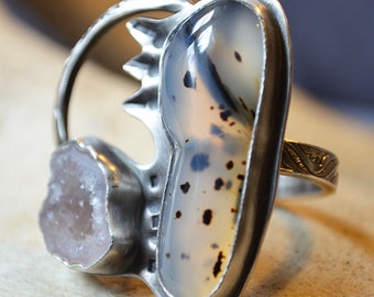 May include: A silver ring with a large, oval-shaped agate stone set in the center. The agate is a light gray color with black and white markings. A smaller, clear quartz stone is set on the side of the ring.