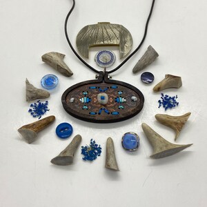 Könnte beinhalten: Ein Holzanhänger mit blauer und weißer Stickerei, umgeben von verschiedenen blauen und weißen Knöpfen und kleinen Geweihstücken. Der Anhänger hängt an einer silbernen Kette.