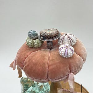 May include: A pink velvet pincushion with a silver ring and several sea shells on top. The ring features a large iridescent shell with a copper wire design.
