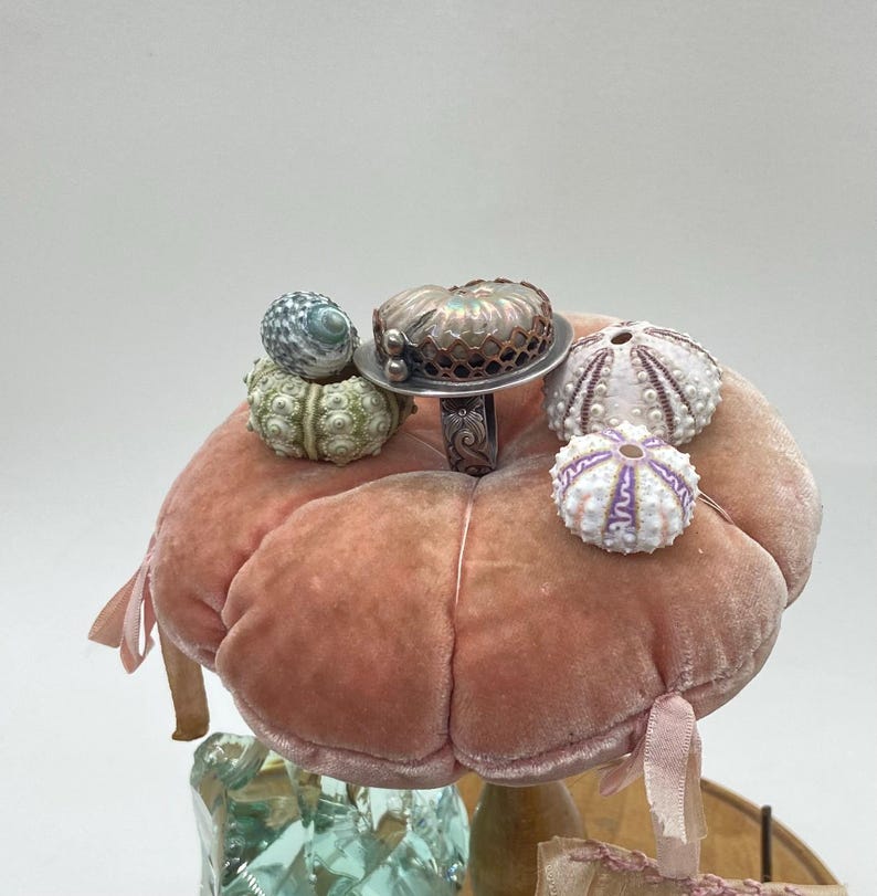 May include: A pink velvet pincushion with a silver ring and several sea shells on top. The ring features a large iridescent shell with a copper wire design.
