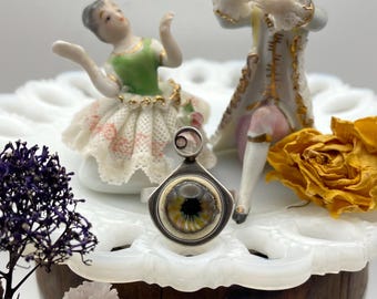 May include: A silver ring featuring a detailed eye design with yellow, green, and black iris. The ring is set against a backdrop of vintage figurines and dried flowers, creating a unique and artistic composition.
