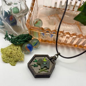 May include: A wooden pendant necklace with a dark brown hexagonal frame. The pendant features a green dog design with green accents and stitching. The necklace is on a black cord, with marbles and decorative moss in the background.