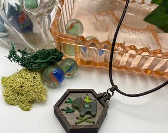 May include: A wooden pendant necklace with a dark brown hexagonal frame. The pendant features a green dog design with green accents and stitching. The necklace is on a black cord, with marbles and decorative moss in the background.