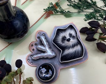May include: A handcrafted brooch featuring a raccoon design with black and white fur. The brooch has a copper-colored base with a silver-colored accent and a small, round, black glass element with a white floral design. The brooch is a unique piece of jewelry.