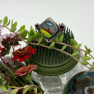 May include: A silver ring with a square, iridescent stone, accented by two small red gemstones. The ring is displayed on a green floral frog, surrounded by fresh flowers and greenery. The stone displays colors of blue, green, and yellow.