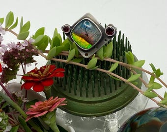 May include: A silver ring with a square, iridescent stone, accented by two small red gemstones. The ring is displayed on a green floral frog, surrounded by fresh flowers and greenery. The stone displays colors of blue, green, and yellow.