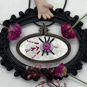 May include: A silver and white embroidered eye with pink accents, framed in a black oval frame. The eye is surrounded by pink flowers.