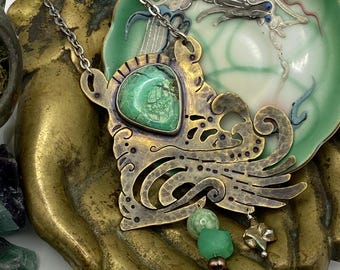 May include: A handcrafted brass pendant necklace with a large, irregularly shaped green stone. The pendant has an ornate, openwork design with a small charm and green beads. The necklace rests on a brass hand-shaped dish.