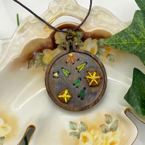 May include: A handcrafted pendant necklace featuring a small, round wooden embroidery hoop. The pendant showcases a stitched design with yellow, green, and orange thread. The necklace has a brown leather cord and a metal clasp.