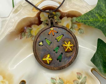May include: A handcrafted pendant necklace featuring a small, round wooden embroidery hoop. The pendant showcases a stitched design with yellow, green, and orange thread. The necklace has a brown leather cord and a metal clasp.