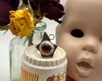 May include: A star-shaped pendant with a red eye design. The pendant is silver-toned and has a small loop at the top. The eye has a red iris and a white sclera. The pendant is displayed on a white and orange ceramic object.