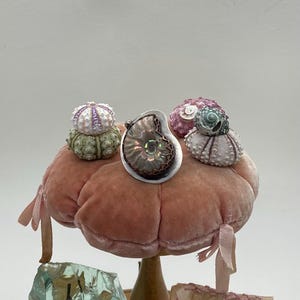 May include: A silver pendant featuring a fossilized ammonite with iridescent colors, surrounded by three small sea urchin shells on a pink velvet pincushion.
