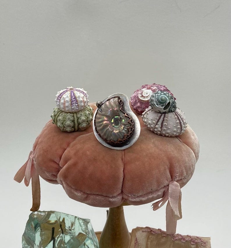 May include: A silver pendant featuring a fossilized ammonite with iridescent colors, surrounded by three small sea urchin shells on a pink velvet pincushion.