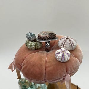 May include: A silver ring with a copper and agate stone set on a pink velvet cushion. The ring is surrounded by several small sea urchin shells.