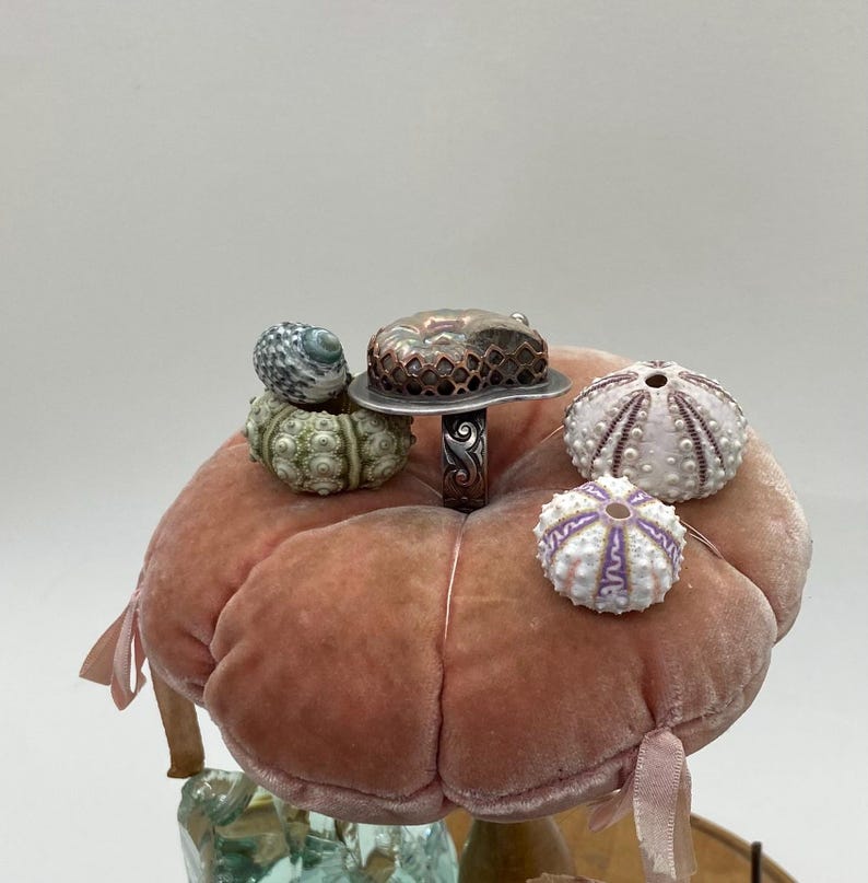May include: A silver ring with a copper and agate stone set on a pink velvet cushion. The ring is surrounded by several small sea urchin shells.