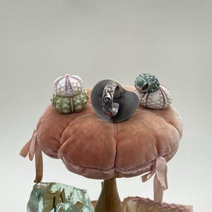 May include: A silver ring with an intricate design sits on a pink velvet cushion, surrounded by three small white and green sea urchins.