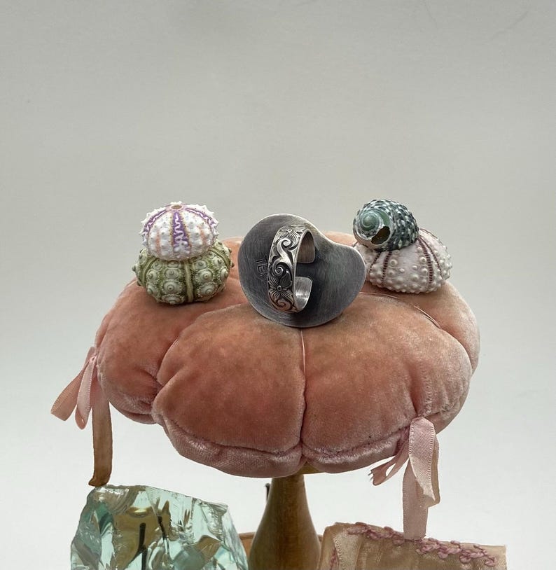 May include: A silver ring with an intricate design sits on a pink velvet cushion, surrounded by three small white and green sea urchins.