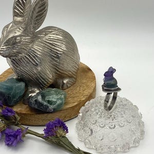 May include: A silver ring with a purple and green fluorite rabbit charm. The ring is sitting on a glass dish with a lace pattern.