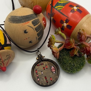 May include: A wooden pendant necklace with a dark brown frame and embroidered red and pink details. The necklace is displayed with several wooden dolls, including a red and black painted doll and a doll with a painted face.