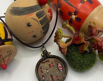 May include: A wooden pendant necklace with a dark brown frame and embroidered red and pink details. The necklace is displayed with several wooden dolls, including a red and black painted doll and a doll with a painted face.