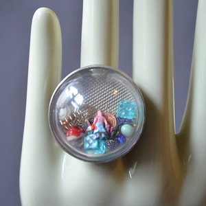 May include: A clear glass ring with a silver band and a variety of colorful beads and trinkets inside. The ring is on a white mannequin hand.