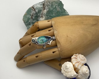 May include: A silver ring with a teardrop-shaped opal stone set in a silver band. The opal is a mix of blue, green, and black colors. The ring is on a wooden hand model.