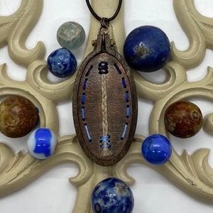 May include: A brown leather oval pendant with blue embroidery, hanging from a black cord. The pendant is surrounded by blue and brown marbles.