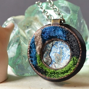 May include: A round copper pendant necklace with a map design in the center. The map is surrounded by layers of blue, green, and brown embroidery.