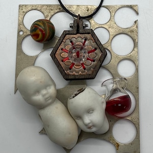 May include: A hexagonal pendant necklace with a red and copper design, a small porcelain doll head, a porcelain doll body, a small glass bottle with red liquid, and a red, yellow, and green marble on a metal background.