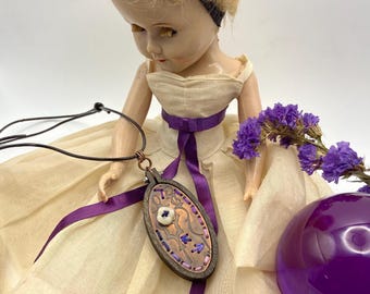 May include: A vintage doll wearing a cream-colored dress with a purple ribbon. A handcrafted pendant with copper and wood accents hangs from a black cord. The pendant features a white button and purple embroidery.