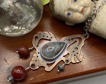 May include: A handcrafted copper pendant necklace with a teardrop-shaped stone and red bead accents. The pendant has an intricate, openwork design. The necklace is displayed on a wooden surface with a vintage doll head and a glass sphere.