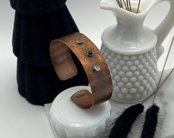May include: A copper cuff bracelet with an etched floral design and three black and white gemstones. The bracelet is displayed on a white surface with black feathers.