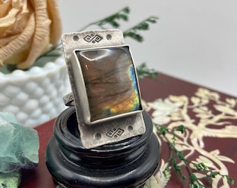 May include: A silver ring with a rectangular labradorite stone. The stone has a mix of colors, including blue, yellow, and brown. The ring has a decorative design on the band and is displayed on a small black stand.