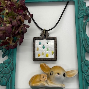 May include: A brown wooden frame with a white background and colorful embroidery. The frame is hanging from a black cord and is attached to a small ceramic deer figurine.