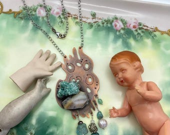 May include: A copper-colored pendant necklace with a silver chain. The pendant features a cluster of green crystals and a stone, with dangling beads in shades of blue, gray, and pearl. The necklace is displayed on a floral-patterned tray.