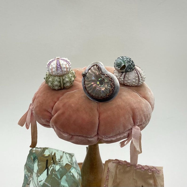 May include: Three small sea shells on a pink velvet pincushion. The shells are a white and green sea urchin, a green and white sea snail, and a iridescent abalone shell.