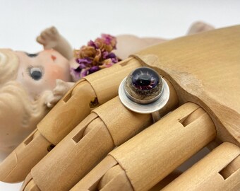 May include: A silver ring with a large, round, dark purple and black stone. The ring is set on a silver base and is displayed on a wooden hand. The ring's stone has a unique, textured appearance.