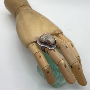 May include: A silver ring with a copper and iridescent ammonite stone set in a textured bezel. The ring is on a wooden hand model.