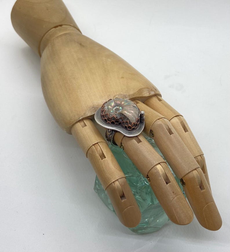 May include: A silver ring with a copper and iridescent ammonite stone set in a textured bezel. The ring is on a wooden hand model.