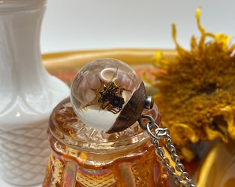 May include: A clear spherical pendant containing a dark, spiky organic element, set into a metallic copper-toned cap with a small embedded bead. The pendant is attached to a silver-toned metal chain, showcasing a detailed, textured design.