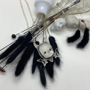 May include: A silver necklace with a pendant featuring a porcelain doll's face framed by a skull-like design. The pendant is adorned with black gemstones and hangs from a silver chain. The necklace is surrounded by black feathers.