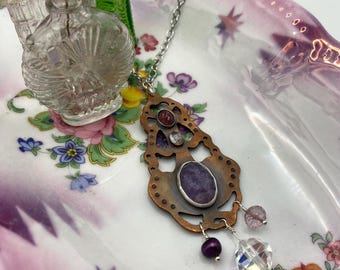 May include: A copper-toned pendant necklace with a silver chain. The pendant features an oval amethyst, a clear crystal, and other gemstones. The necklace is displayed on a floral patterned dish with small glass bottles.