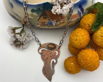 May include: A necklace featuring a copper-toned metal pendant with an organic, asymmetrical shape and etched line details. The pendant is set with a smooth, oval brown stone at the top and a small, rectangular amber-colored bead at the bottom.