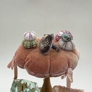May include: A silver ring with a green and brown iridescent stone set in a silver and copper cage design. The ring is displayed on a pink velvet cushion with three small sea urchin shells.