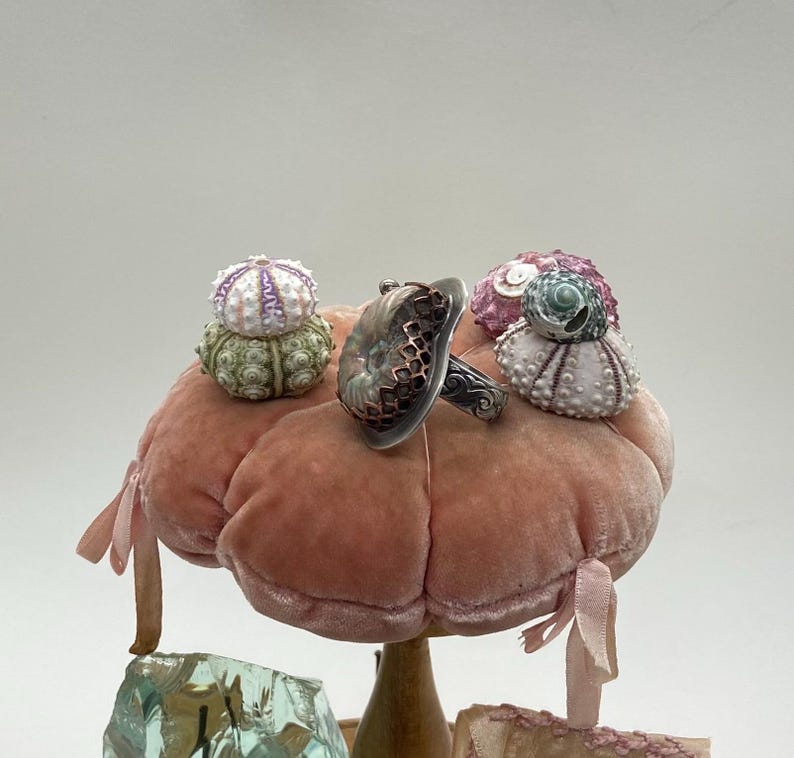 May include: A silver ring with a green and brown iridescent stone set in a silver and copper cage design. The ring is displayed on a pink velvet cushion with three small sea urchin shells.