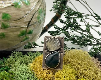 May include: A rectangular silver ring featuring two stones set vertically. The top stone is a translucent, textured, light-colored piece, while the bottom stone is a smooth, dark green, teardrop-shaped gem. The silver band has etched markings.
