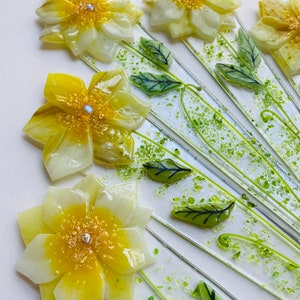 May include: A glass art piece featuring a cluster of yellow and white flowers with green leaves on a white background. The flowers are made of glass and have a textured surface. The leaves are also made of glass and have a green color.