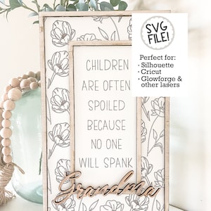 Könnte beinhalten: Ein weißes Holzschild mit einem Blumenmuster und dem Text "CHILDREN ARE OFTEN SPOILED BECAUSE NO ONE WILL SPANK GRANDMA". Das Schild ist in einem weißen Rahmen.