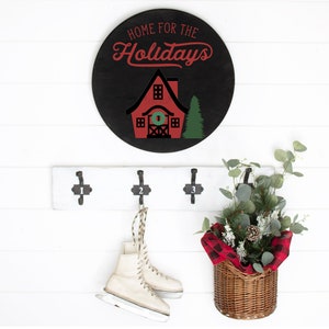 May include: A black and red round wooden sign with the text "Home for the Holidays" and a red house with a green wreath on the door. The sign is hanging on a white wall with a white wooden coat rack with three hooks. A pair of white ice skates are hanging on one of the hooks. A wicker basket with a red and black plaid fabric liner is hanging on another hook. The basket is filled with green eucalyptus branches and a pine cone.