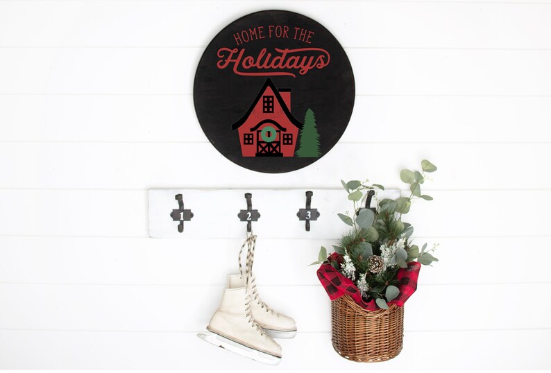 May include: A black and red round wooden sign with the text "Home for the Holidays" and a red house with a green wreath on the door. The sign is hanging on a white wall with a white wooden coat rack with three hooks. A pair of white ice skates are hanging on one of the hooks. A wicker basket with a red and black plaid fabric liner is hanging on another hook. The basket is filled with green eucalyptus branches and a pine cone.